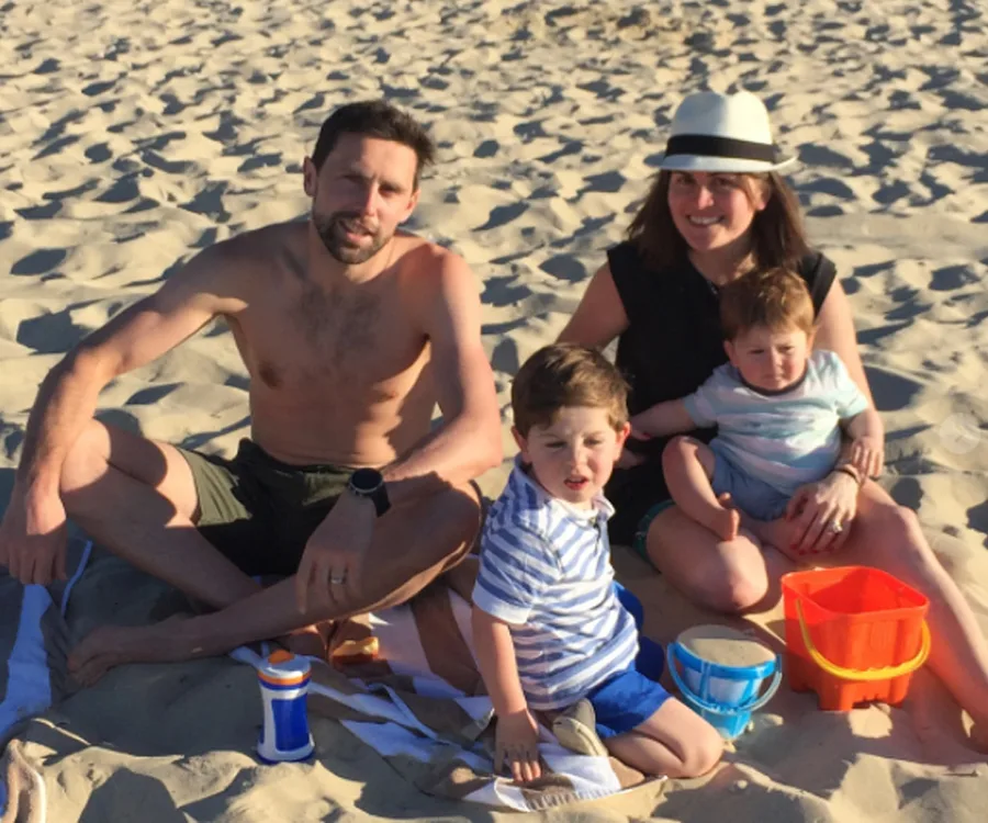 A family of four sitting on a sandy beach; a man, woman, and two young children with sand toys.