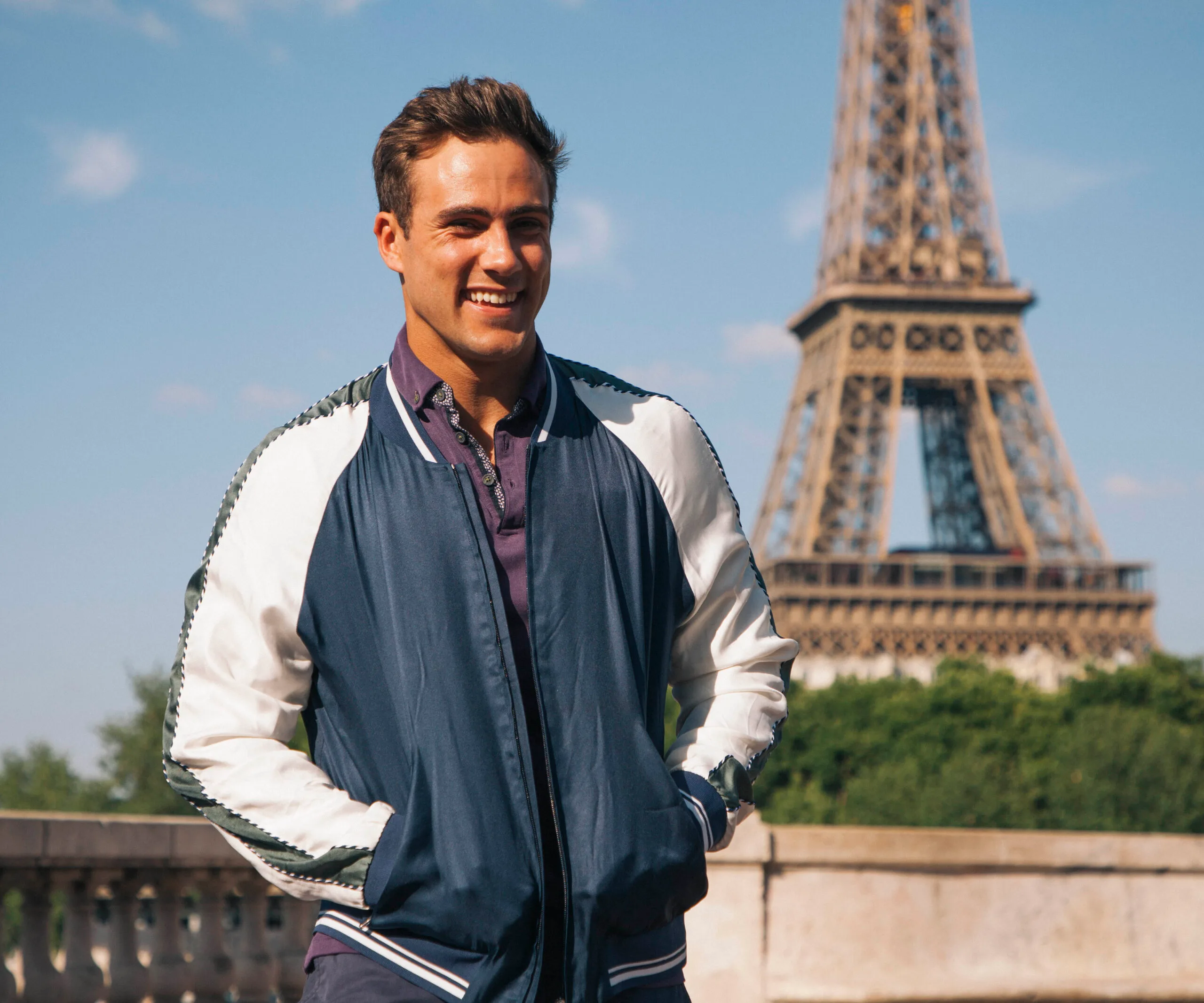 Man smiling in front of the Eiffel Tower, wearing a blue and white jacket.