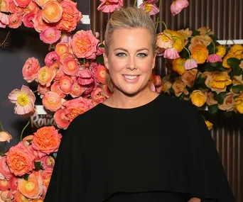 Smiling woman in a black dress stands in front of colorful flowers.