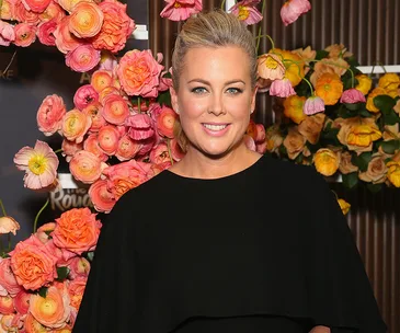 Smiling woman in a black dress stands in front of colorful flowers.
