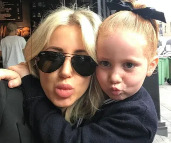 Adult and child posing with pouty lips; adult in sunglasses, child with red hair and bow, outdoors.