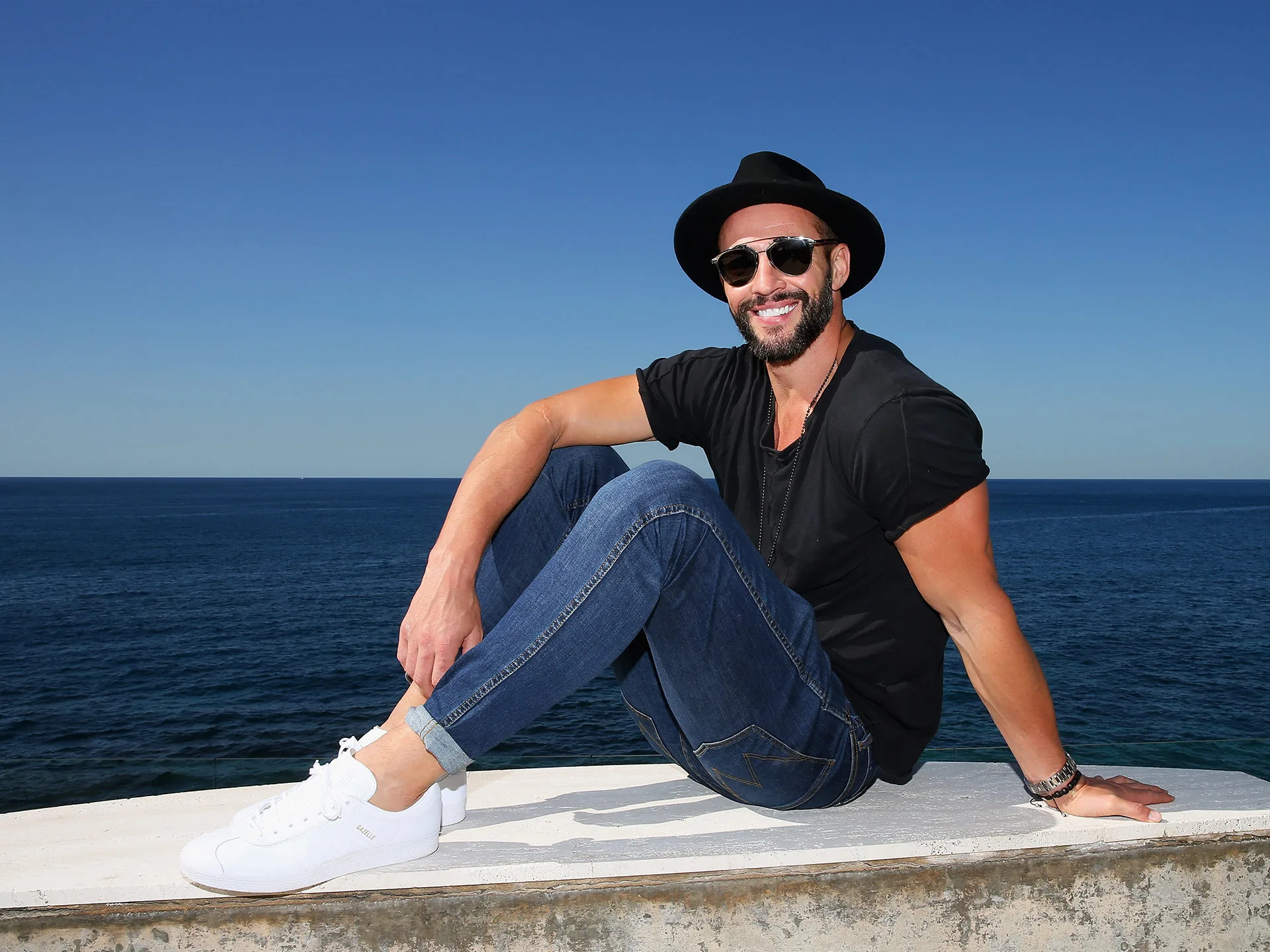 Man in a hat and sunglasses smiles while sitting by the ocean on a sunny day.