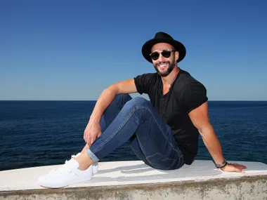 Man in a hat and sunglasses smiles while sitting by the ocean on a sunny day.