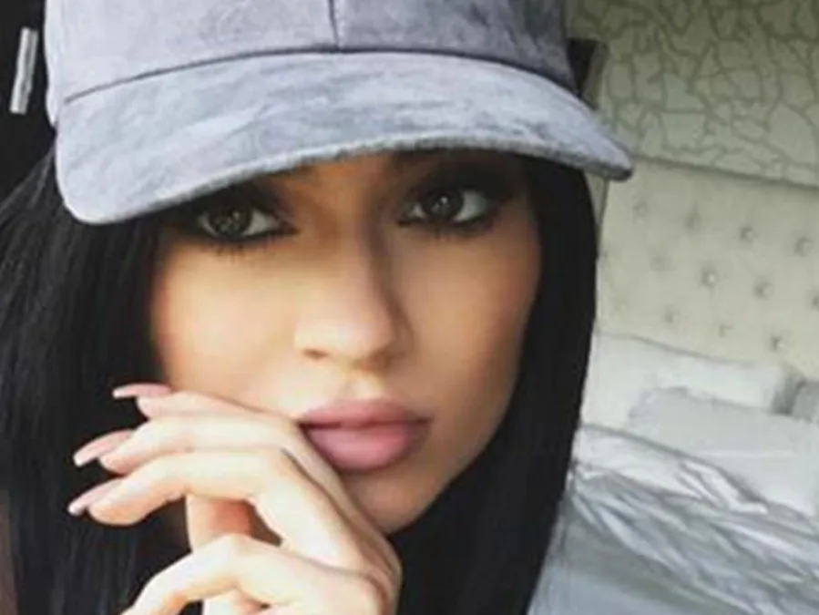 Young woman with dark hair wearing a grey cap, posed indoors, resting her chin on her hand, with a neutral expression.