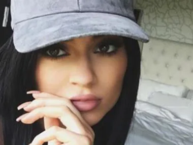 Young woman with dark hair wearing a grey cap, posed indoors, resting her chin on her hand, with a neutral expression.