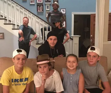 Group of people posing in a living room with stairs, one wearing a "Modern Family" shirt, others in casual wear.