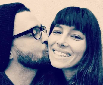 "Man in beanie and glasses kisses smiling woman with bangs, showing affection and happiness."