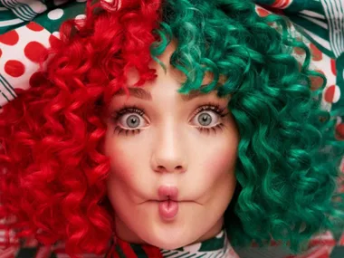 A person with wide eyes and curly, half red, half green hair puckers their lips in a playful expression.