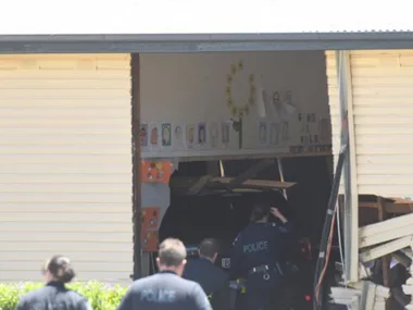 Police officers inspecting vehicle crashed into building; classroom art visible inside.