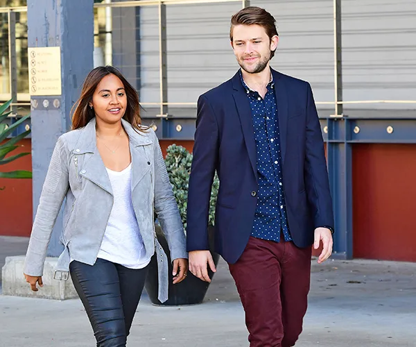 Jessica Mauboy and a male companion walk together, she in a gray jacket and he in a navy blazer, outdoors.