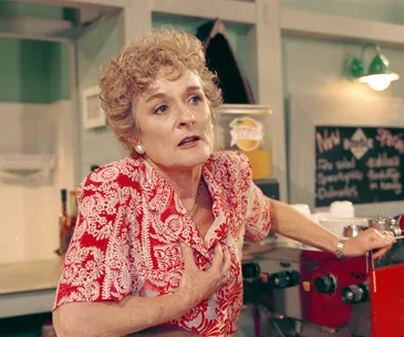 Elderly woman in a red floral blouse, clutching her chest in distress in a kitchen setting.