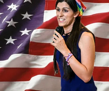 A woman speaking with a microphone in front of a large American flag backdrop, wearing a colorful headscarf.