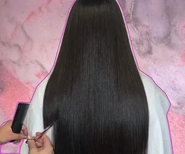 A stylist cuts long dark hair into a geometric triangle shape, using a comb and scissors, against a pink marble background.