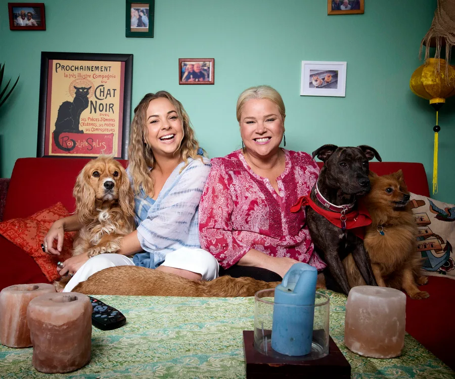 Two women on a sofa with three dogs, surrounded by framed photos and art in a cozy living room setting.