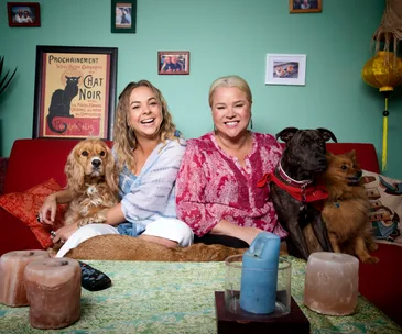 Two women on a sofa with three dogs, surrounded by framed photos and art in a cozy living room setting.