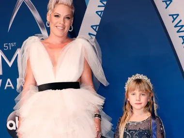 A person in a white dress and short blonde hair stands next to a young girl in a sparkly dress and headband on a blue carpet.