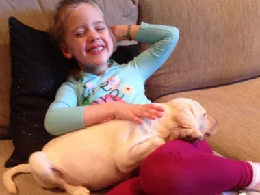 A smiling child reclines on a couch, gently holding a sleeping puppy on their lap.