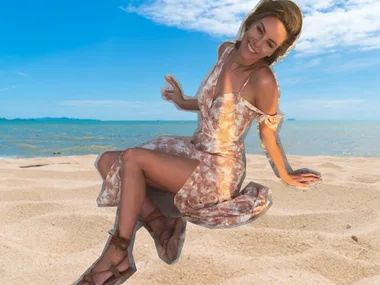 Woman in a floral dress sitting on a sunny beach with a joyful expression against a clear blue sky and ocean backdrop.