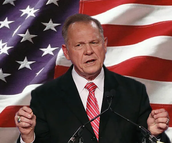 A man in a suit and red tie speaks in front of an American flag backdrop.