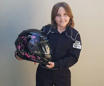 A young person in a racing suit holding a black helmet with pink designs against a plain background.