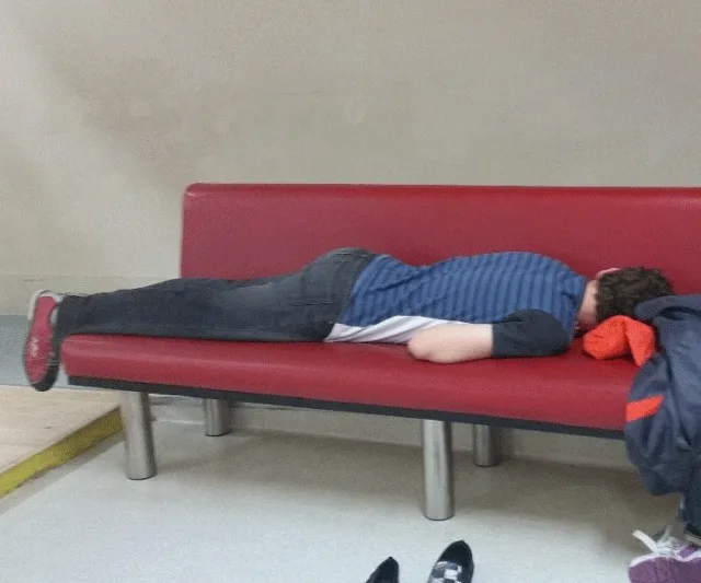 Person sleeping face down on a red bench in casual attire, shoes off, head on a jacket.