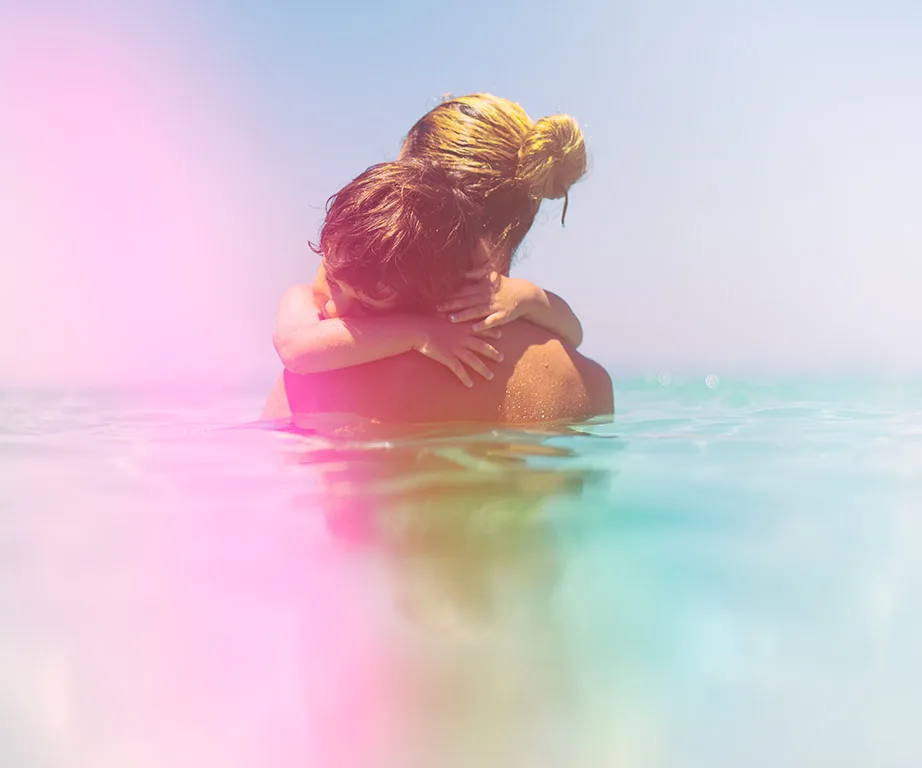 A woman and child embrace in a pastel-tinted ocean, with the child's arms around the woman's neck.