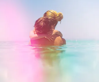 A woman and child embrace in a pastel-tinted ocean, with the child's arms around the woman's neck.