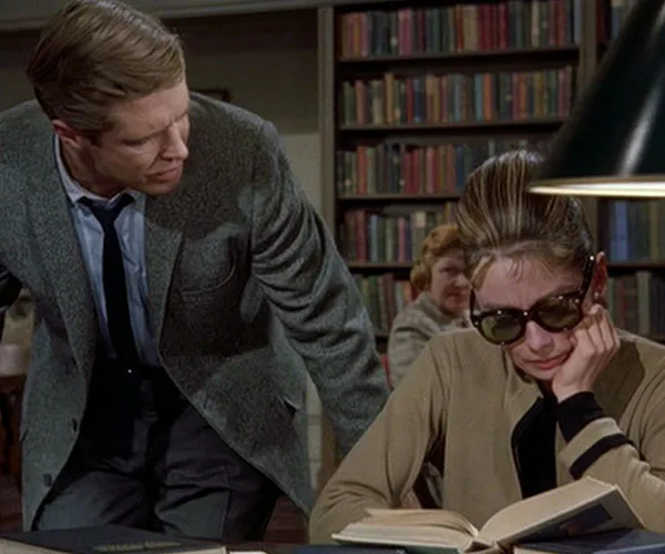 Man in a suit leans over a woman wearing sunglasses, both in a library setting with bookshelves in the background.