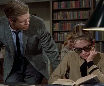 Man in a suit leans over a woman wearing sunglasses, both in a library setting with bookshelves in the background.