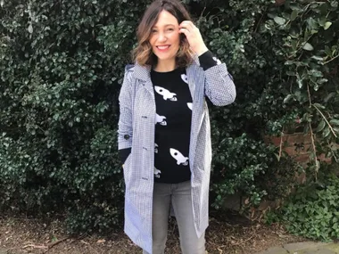 Woman smiling in black rocket sweater and checkered coat, standing in front of a leafy green bush.
