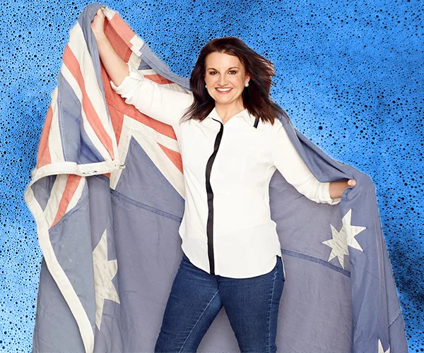 Australian politician Jacqui Lambie stands smiling, holding an Australian flag against a blue background.