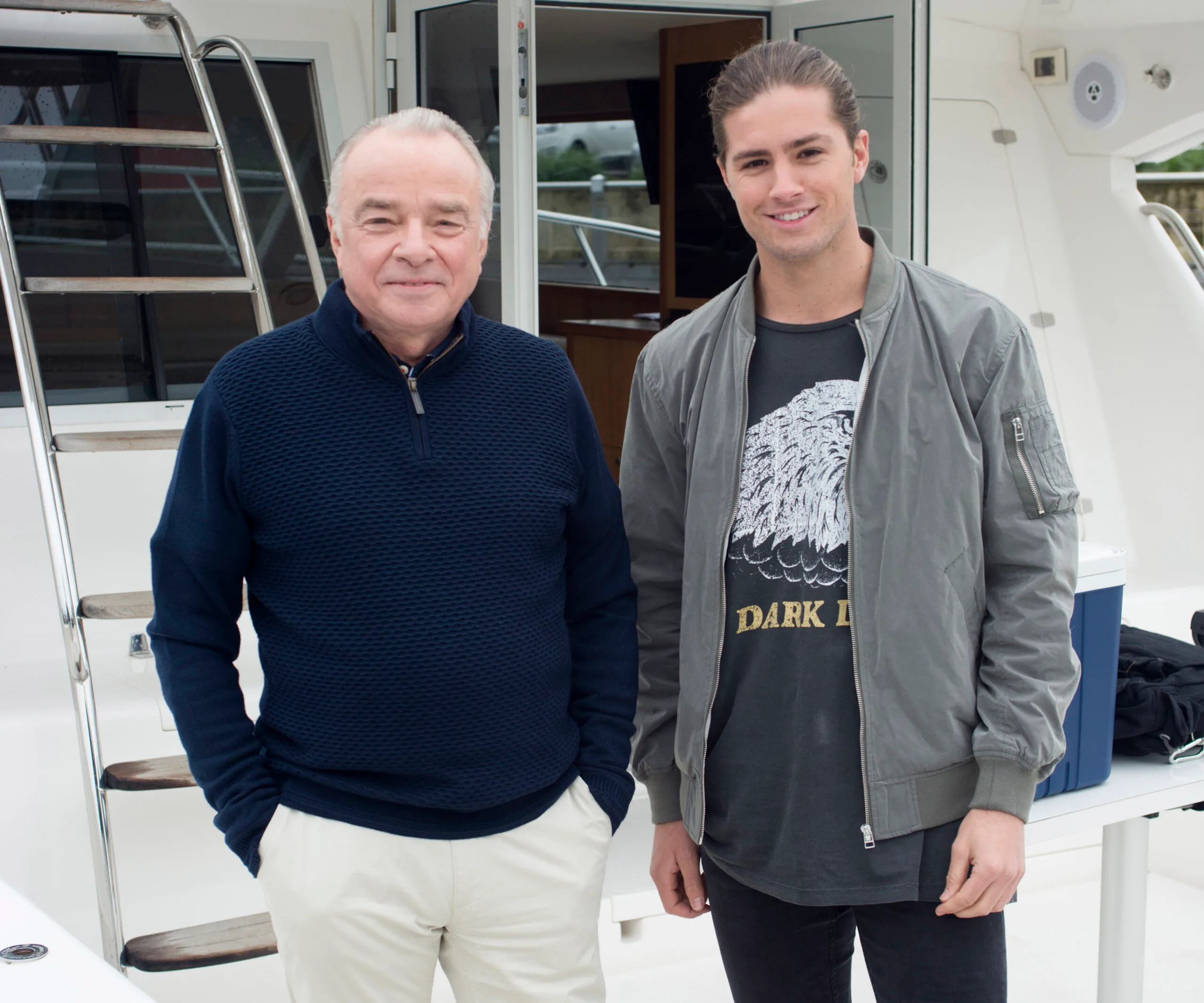 Two men smiling on a boat, with one wearing a navy sweater and the other in a gray jacket and graphic t-shirt.