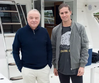 Two men smiling on a boat, with one wearing a navy sweater and the other in a gray jacket and graphic t-shirt.