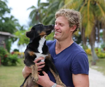 Man smiling while holding a dog outdoors with palm trees in the background.