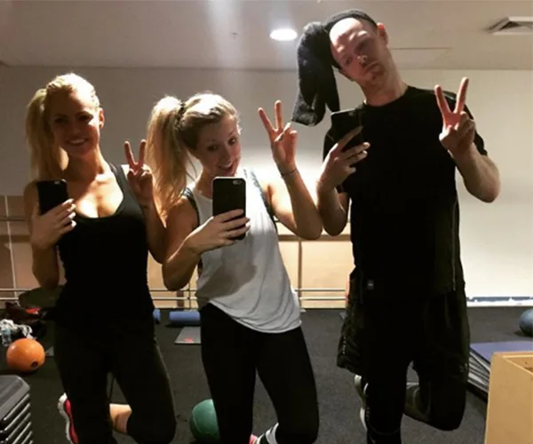 Three people posing in a gym, taking a mirror selfie while holding up peace signs.