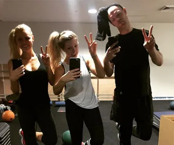 Three people posing in a gym, taking a mirror selfie while holding up peace signs.