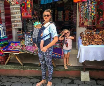 Mother with baby in carrier and child posing outside colorful textile shop.