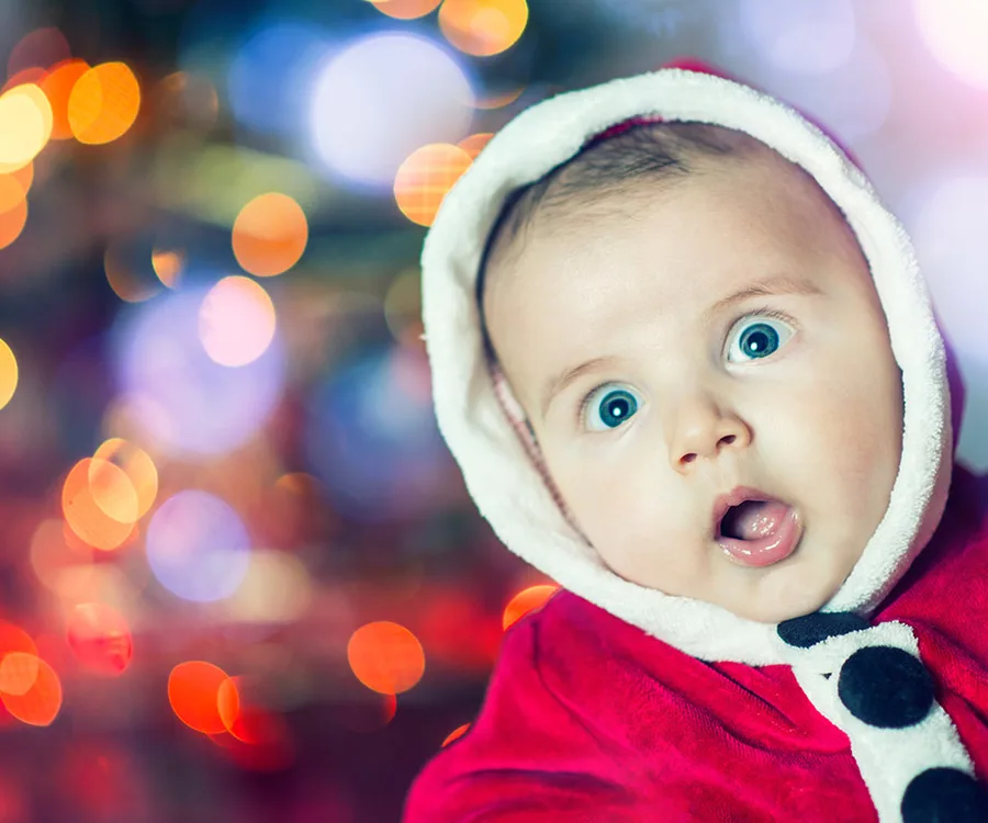 A baby in a red Santa suit looks surprised, with colorful holiday lights blurred in the background.