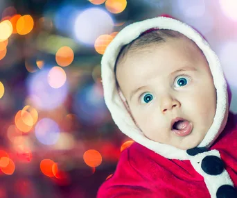 A baby in a red Santa suit looks surprised, with colorful holiday lights blurred in the background.