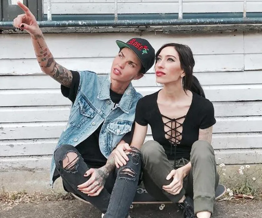 Two people sitting on a skateboard, one pointing into the distance, in front of a white wooden building.