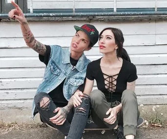 Two people sitting on a skateboard, one pointing into the distance, in front of a white wooden building.