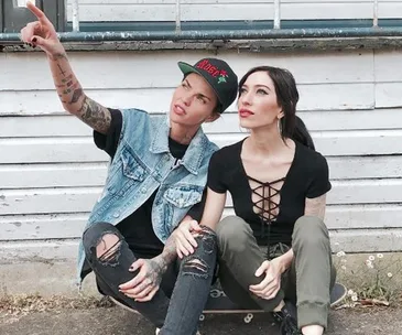 Two people sitting on a skateboard, one pointing into the distance, in front of a white wooden building.