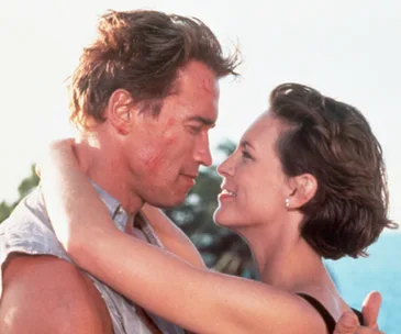 A man and woman embrace closely, gazing into each other's eyes, against a bright, outdoor background.
