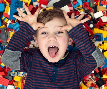 Child playing on pile of colorful LEGO bricks, making a playful face with tongue out and hands on head.
