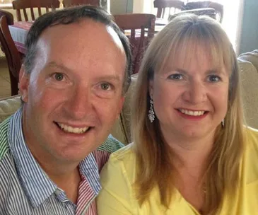 A smiling man and woman sitting together indoors, with chairs and tables in the background.
