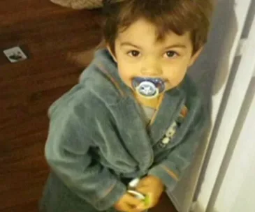 A toddler in a robe with a pacifier looks up at the camera, standing indoors on a hardwood floor.