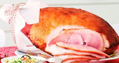 Roasted Christmas ham with slices, garnished with festive ribbon, served on a platter with a side dish.