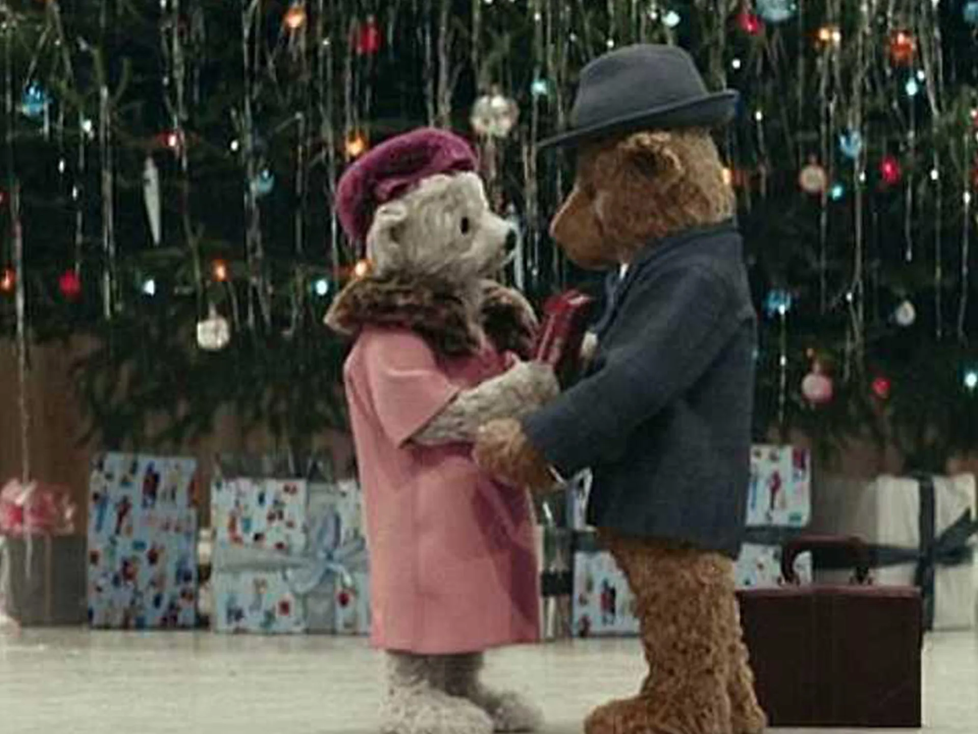 Teddy bears embrace by a Christmas tree, wearing festive clothes, part of Heathrow's Christmas ad scene.