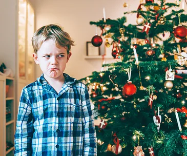 Child in pajamas frowning near a decorated Christmas tree.