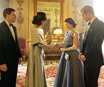 A formal meeting between two women in elegant gowns, with men in suits, in a richly decorated room with a large mirror.